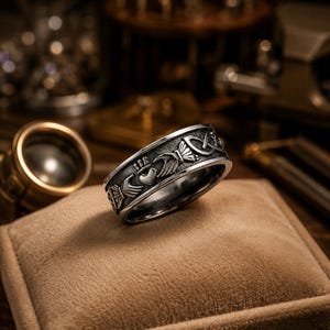 Puede incluir: Un anillo de metal plateado con un diseño Claddagh en relieve que muestra dos manos sosteniendo un corazón coronado, seguido de un intrincado patrón de nudo celta. La banda tiene un acabado oscuro y oxidado en las áreas hundidas del diseño.