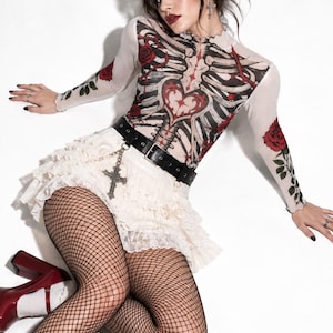 May include: A long-sleeve sheer white top featuring a graphic print of a human rib cage, a central heart, and red roses. The outfit includes a cream-coloured tiered lace mini skirt, a black leather belt with a silver cross charm, and black fishnet tights.