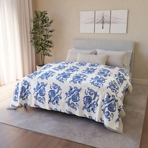 May include: A white bed blanket featuring a repeating blue pattern of stylised dragon-like creatures and floral motifs. The blanket is draped over a bed with a grey headboard, light grey pillows, and a grey rug on a wooden floor in a room.