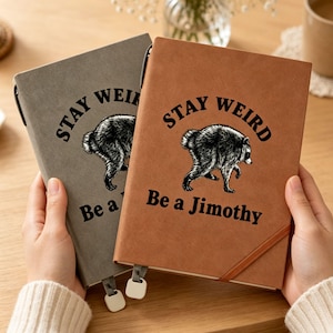 May include: Two rectangular notebooks, one gray and one brown, each featuring a black graphic of a raccoon and the text "STAY WEIRD Be a Jimothy" printed on the front cover. Both notebooks include a matching elastic closure band and a ribbon bookmark.