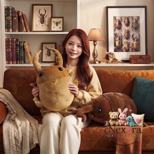 May include: A large, tan, plush beetle toy with prominent mandibles and dark eyes, held by a person. Beside it sits a larger brown beetle plush, a small tan bear, a pink rabbit, a blue dinosaur, and a small square pillow with a pink heart.