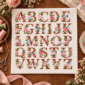 May include: A square white fabric piece featuring a cross-stitch alphabet from A to Z. Each brown letter is intricately decorated with small pink, blue, and white floral patterns and green leaves, arranged in five rows on the canvas.