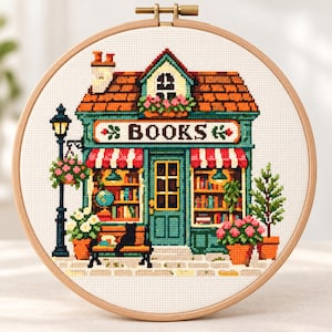 May include: A circular wooden embroidery hoop containing a cross-stitch pattern of a bookstore with a teal facade, orange roof, and red-and-white striped awnings. The storefront features the word BOOKS, a black cat on a bench, and potted flowers.