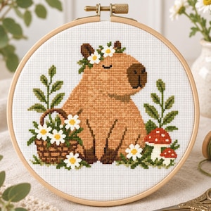 May include: A cross-stitch embroidery project in a round wooden hoop featuring a tan capybara with a white flower crown. The design includes a brown woven basket filled with white daisies, green leafy sprigs, and two red and white mushrooms.