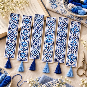 May include: Six rectangular bookmarks featuring intricate cross-stitch embroidery in various shades of blue and white. Each bookmark displays a unique geometric or floral pattern and is finished with a matching blue thread tassel at the bottom.