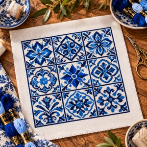 Op de afbeelding: Een vierkant stuk witte stof met een kruissteekpatroon bestaande uit negen afzonderlijke tegels. Elke tegel toont een uniek geometrisch of bloemmotief in verschillende blauwtinten, wat samen een samenhangend rasterontwerp vormt.
