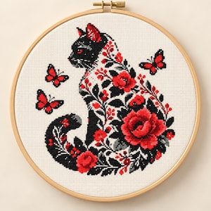 May include: A circular wooden embroidery hoop containing a cross-stitch pattern of a black cat silhouette filled with intricate red roses, black leaves, and small red berries, accompanied by two red butterflies on a white fabric background.