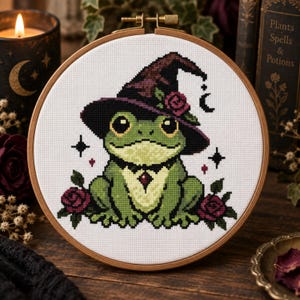 May include: A cross-stitch embroidery in a round wooden hoop featuring a green frog wearing a dark purple witch hat adorned with a rose. The frog has a light green belly with a small red gem and is flanked by two dark red roses on a white fabric.