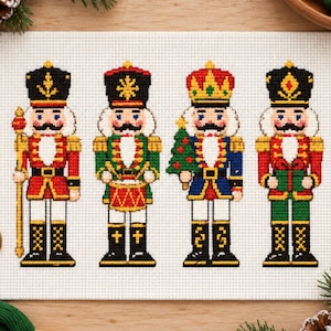 May include: A cross-stitch embroidery on white Aida fabric featuring four nutcracker figures. The figures wear red, green, and blue uniforms with gold accents, black boots, and hats. One holds a staff, one a drum, and one a small Christmas tree.
