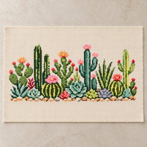 Op de afbeelding: Een rechthoekig kruissteekborduurwerk op crèmekleurige stof met een verscheidenheid aan groene cactussen en vetplanten. De planten zijn versierd met kleine roze, oranje en rode bloemen, gerangschikt in een rij op een basis van kleine bruine steentjes.