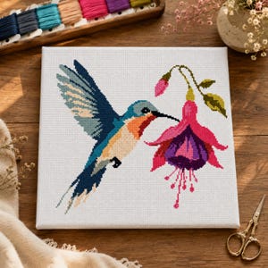 Puede incluir: Un bordado de punto de cruz cuadrado sobre tela blanca que muestra un colibrí con plumaje azul, naranja y blanco volando cerca de una flor de fucsia de colores rosa intenso y morado. El diseño está realizado en estilo píxel sobre fondo blanco.