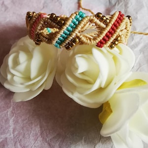 May include: A macrame bracelet featuring a woven beige cord pattern accented with vibrant turquoise, red, and pink thread sections. The design is embellished with small, metallic bronze-colored beads integrated throughout the braided structure.