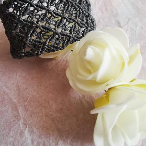 May include: A dark grey macrame bracelet featuring an intricate, repeating diamond-shaped knot pattern. The bracelet is constructed from thin, cord-like material and includes adjustable strings at the ends for a secure fit around the wrist.