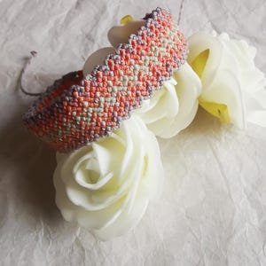 Könnte beinhalten: Ein gewebtes Makramee-Armband mit einem sich wiederholenden Zickzackmuster in Koralle, Hellgrün und Grau. Das Armband hat eine strukturierte, geknotete Oberfläche mit dünnen grauen Kordeln an den Enden für einen verstellbaren Verschluss.