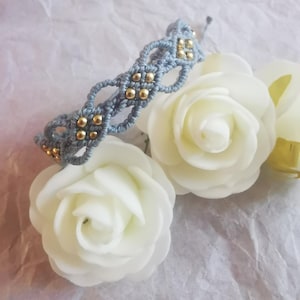 May include: A light blue macrame bracelet featuring a woven cord pattern with small, round, metallic gold-colored beads integrated throughout the design. The bracelet is positioned above two white artificial rose flowers.