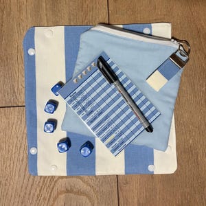 Könnte beinhalten: Eine blau-weiß gestreifte Stoffmatte mit Druckknöpfen, ein hellblaues Reißverschlusstäschchen mit passendem Schlüsselanhänger, fünf blaue Würfel und ein Punkteblock mit schwarzem Stift. Der Block enthält Spalten für Namen und Spielkategorien.