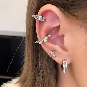 Puede incluir: Un conjunto de joyería plateada para la oreja que incluye un pendiente de clip con púas en el cartílago superior, un aro con púas en la concha, un pequeño pendiente de botón con forma de letra K con piedras claras y un aro pequeño con púas.