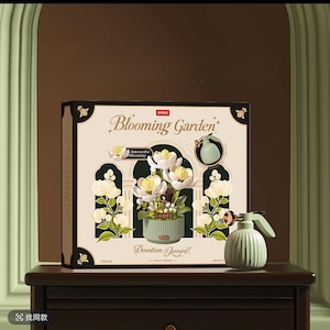 Puede incluir: Una caja de producto cuadrada para un set de construcción Blooming Garden con flores de jazmín blancas en una maceta verde acanalada. La caja muestra el texto Devotion of Jasmine e Interactive Blooming. Un pequeño pulverizador verde acanalado se sitúa al lado.