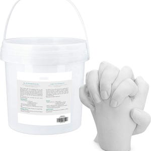 May include: A white plastic bucket with a handle and a label containing product information, accompanied by a white 3D plaster cast sculpture of two hands clasped together. The label includes text detailing the contents of the casting kit.