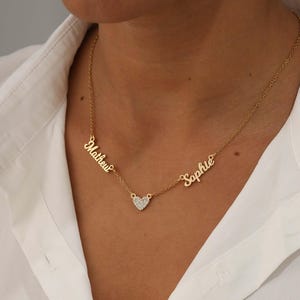 Puede incluir: Un collar de tono dorado con una cadena fina y dos colgantes con los nombres Mathew y Sophie en letra cursiva, unidos por un pequeño dije central en forma de corazón incrustado con diminutas piedras transparentes sobre una camisa blanca.