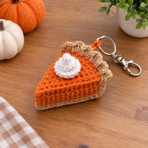Puede incluir: Un llavero de ganchillo con forma de porción de tarta de calabaza con corteza color canela, relleno naranja y un copete de nata montada blanca encima. La porción de tarta está unida a una anilla de metal plateado y un cierre de mosquetón.