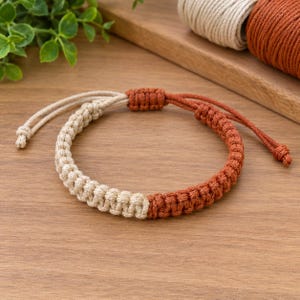 May include: A macrame bracelet featuring a two-tone design with one half in cream and the other half in terracotta orange. The bracelet is constructed with a series of square knots and includes an adjustable sliding knot closure with loose cord ends.