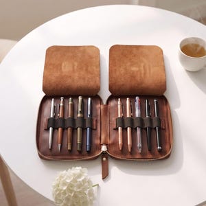 以下が含まれることがあります： ファスナー付きの茶色のレザー製ペンケース。平らに開くと、10本の様々なペンを収納できる伸縮性のあるループを備えた2つの内部セクションが現れます。滑らかなレザーの外装と柔らかい内装の裏地が特徴で、筆記具を整理して収納できます。