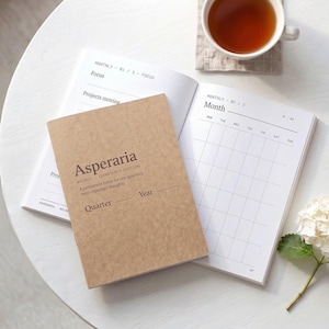 Puede incluir: Una agenda con cubierta de papel color canela titulada Asperaria Weekly Quarterly Edition con un espacio en blanco para el trimestre y el año. La agenda abierta muestra una cuadrícula de calendario mensual y páginas de seguimiento de proyectos.