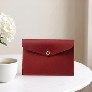Könnte beinhalten: Eine rechteckige, rote Ledertasche im Umschlagstil mit einer spitzen Klappe und einem einzelnen, mittig platzierten silberfarbenen Metalldruckknopf. Die Tasche hat eine glatte, matte Oberfläche und sichtbare Nähte an den unteren und seitlichen Kanten.