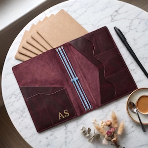 May include: A deep burgundy leather traveler's notebook cover featuring three light blue elastic cords for securing inserts, internal pockets with wavy edges, and the gold-embossed initials AS on the front pocket, accompanied by several tan paper inserts.