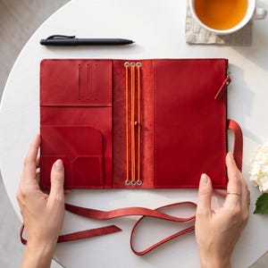 May include: A bright red leather traveler's notebook cover featuring an open interior with multiple card slots, a large pocket, and three vertical orange elastic bands for securing inserts. A matching red leather strap is attached to the side.
