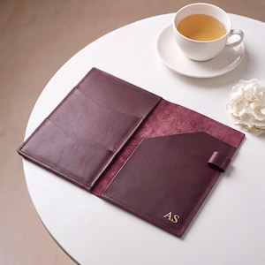 Puede incluir: Una funda de cuero color burdeos oscuro para cuaderno con bolsillos internos, un portalápices y las iniciales AS grabadas en oro en la esquina inferior derecha. La funda está abierta, mostrando un acabado liso y una superficie texturizada.