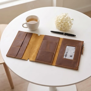 Puede incluir: Una cartera de cuero marrón de tres pliegues con múltiples ranuras para tarjetas, una ventana transparente para identificación y una cremallera lateral. La cartera está abierta y extendida, mostrando su textura suave y costuras visibles.