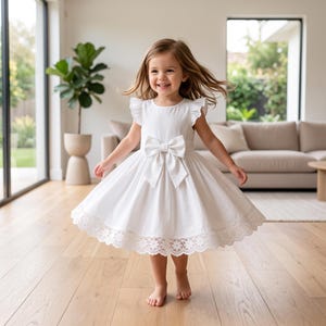 Puede incluir: Un vestido blanco sin mangas con hombros con volantes, un gran lazo central en la cintura y una falda acampanada con un delicado borde de encaje floral a lo largo del dobladillo. El vestido tiene un cuello redondo y un corpiño estructurado.