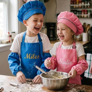 Pode incluir: Um conjunto de chapéu de chef e avental azul com o nome Miles bordado em dourado, e um conjunto de chapéu de chef e avental rosa com o nome Sadie bordado em branco. Ambos incluem mangas protetoras a condizer sobre t-shirts brancas.