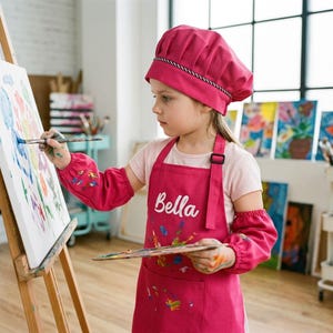Könnte beinhalten: Ein leuchtend pinkfarbenes Schürzen- und Kochmützenset mit dem in weißer Schreibschrift auf die Brust gestickten Namen Bella. Die Schürze hat lange Ärmel mit elastischen Bündchen und eine Vordertasche, alles mit bunten Farbspritzern verziert.