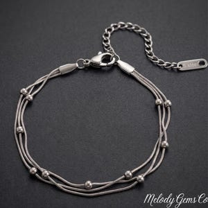 May include: A silver-toned multi-strand bracelet featuring three delicate snake chains adorned with small, evenly spaced round beads. The bracelet includes a lobster claw clasp and an adjustable extender chain with a tag engraved with S.steel.