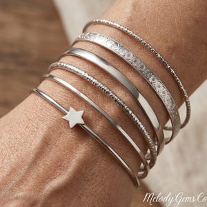 May include: A set of six silver-toned metal bangles worn together. The collection includes a thin textured band, a wider band with an embossed circular pattern, a smooth polished band, a hammered band, a plain thin band, and a thin band with a star.