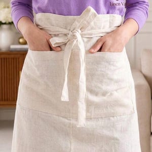 Pode incluir: Um avental de cintura em linho bege com um cós largo, um fecho de laço central e dois bolsos frontais grandes. O tecido tem uma textura de trama natural visível. O logótipo Linen Vastu está impresso no canto superior direito da imagem.