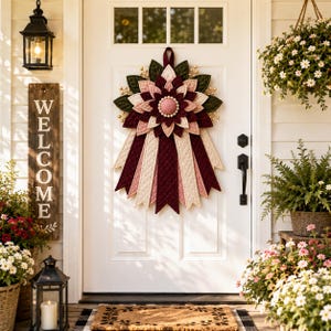 Può includere: Una decorazione da parete in tessuto a forma di grande fiore di dalia con petali stratificati nei toni del bordeaux, rosa antico e crema. Il design presenta un accento circolare centrale con piccole perline bianche e lunghi nastri di tessuto trapuntato.
