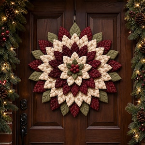 May include: A circular fabric wreath featuring layered, quilted leaf-shaped petals in deep burgundy, cream with a gold snowflake pattern, and sage green. The centre is accented with a cluster of small, round, textured red berries.