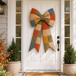 May include: A large, quilted fabric bow decoration featuring a patchwork design of various autumn-toned patterns in shades of orange, olive green, blue, and cream. The bow has two wide loops and two long, pointed tails, centered by a solid olive green band.