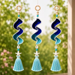 May include: Three hanging ornaments featuring a spiral crochet design in shades of dark blue, teal, and light blue. Each piece includes a wooden ring at the top, wooden and turquoise beads, a white seashell, a white starfish, and a light blue tassel.