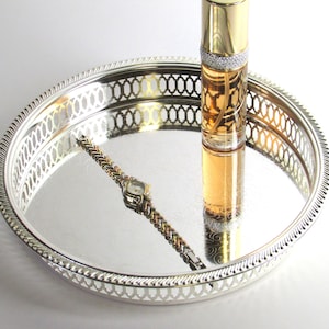 May include: A silver-colored, round tray with a decorative pierced border. A gold-topped perfume bottle with a diamond-like band and a gold and silver watch are on the tray. The perfume bottle contains a light amber liquid.