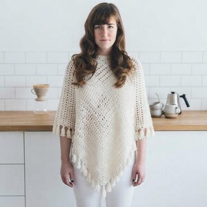 May include: A cream-colored crochet poncho featuring a diamond-patterned knit design with a pointed hemline. The garment is finished with small, matching cream-colored tassels along the bottom edges, creating a textured and layered appearance.