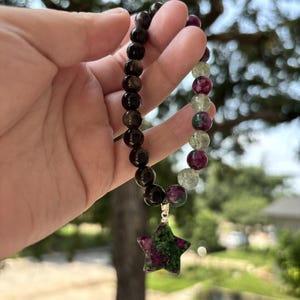 May include: A beaded bracelet featuring a mix of dark brown, translucent light green, and deep magenta spherical beads. A star-shaped pendant made of mottled magenta and green stone hangs from the bracelet, attached by a silver-toned metal bail.