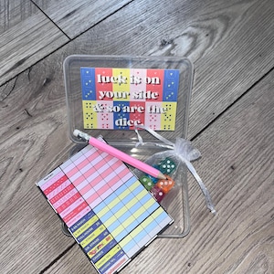 May include: A clear plastic game box containing a score sheet with German text, a pink pencil, and a small white organza bag filled with multi-colored dice. The box lid features a colorful card with the text, luck is on your side & so are the dice.