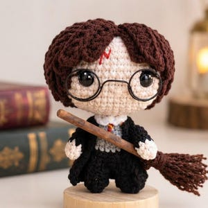 May include: A crocheted doll featuring dark brown hair, a red lightning bolt mark on the forehead, and round black glasses. The doll wears a black robe over a grey vest and white shirt, holding a brown yarn broomstick across the front of the body.