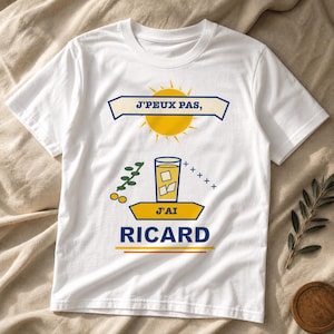 Peut inclure: Un t-shirt blanc à manches courtes avec un imprimé graphique représentant un soleil jaune, un verre de liquide jaune avec des glaçons et le texte J'PEUX PAS, J'AI RICARD. Le motif inclut un brin vert et des petites croix bleues.