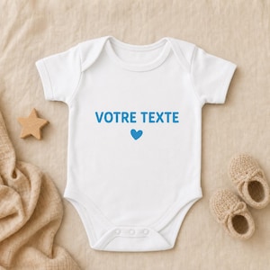 Peut inclure: Un body pour bébé blanc à manches courtes avec le texte bleu "VOTRE TEXTE" imprimé sur la poitrine, placé au-dessus d'une petite icône de cœur bleu. Le vêtement présente une encolure ronde et des boutons-pression à l'entrejambe.